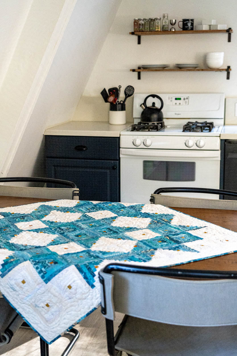 Kitchen with a table covered by a blue and white quilt, shelves with kitchen items, and a stove.